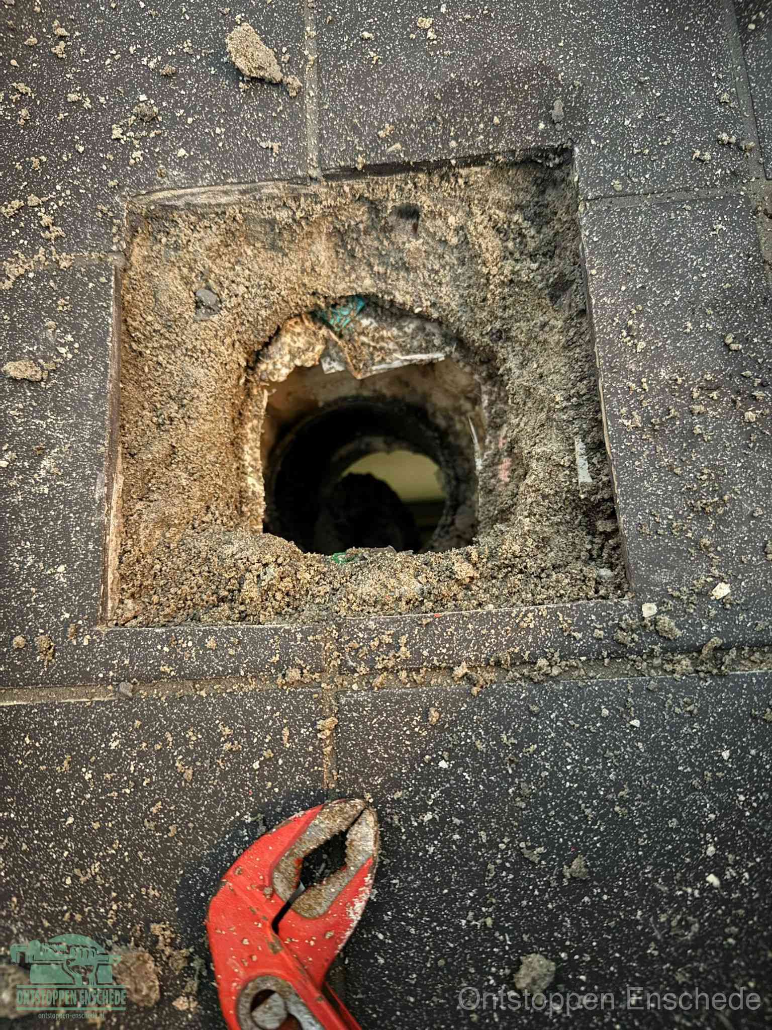 Een geopend rioolputje in een betontegelvloer met losgemaakt rooster en een zichtbare ronde afvoerbuis, het achterhalen waar de afvoerverstopping zit in Enschede