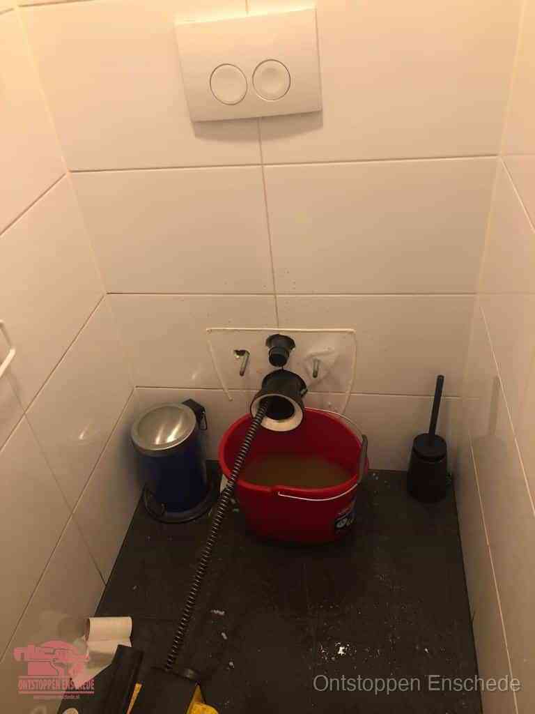Een ontstopte wand-wc met verwijderde toiletpot waar een ontstoppingsveer in de wanduitgang verdwijnt, het achterhalen waar de verstopping zit in Enschede