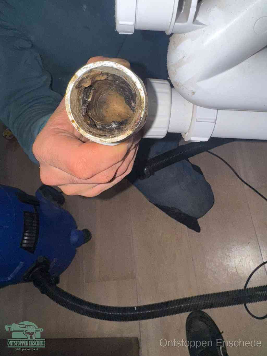 Een hand houdt een losgekoppeld PVC-afvoerbuisstuk vast dat van binnen volledig is dichtgeslibd met vet, vuil en aanslag, de meest voorkomende oorzaak van een verstopte afvoer in Enschede