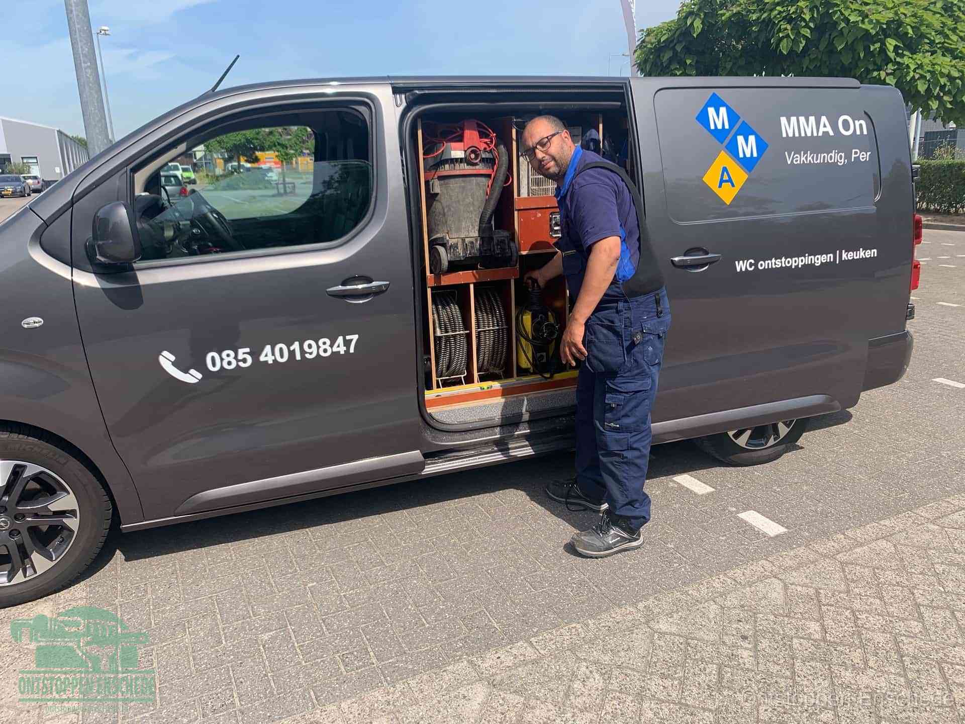 Een monteur bij de open schuifdeur van zijn bedrijfsbus op een parkeerplaats, klaar om de oorzaak van terugkerende afvoerverstoppingen aan te pakken in Enschede