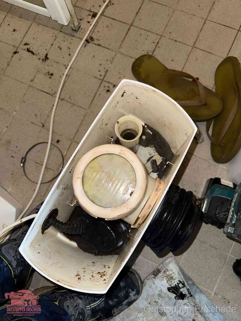 Een gedemonteerde toiletpot op de vloer met ontstoppingsveer en gereedschap ernaast, het verwijderen van een vastzittend voorwerp uit de wc in Enschede