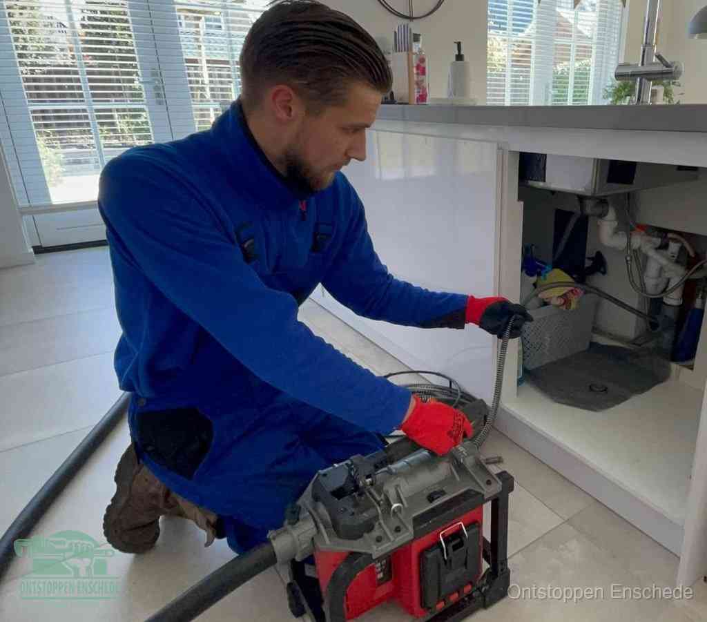 Een monteur met rode handschoenen knielt in een keuken en voert een elektrische ontstoppingsveer in tijdens het professioneel afvoer ontstoppen in Enschede